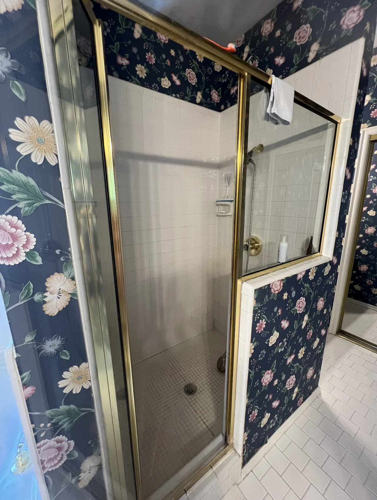 Before: close view of original white-tiled shower with brass-framed glass door and floral wallpaper
